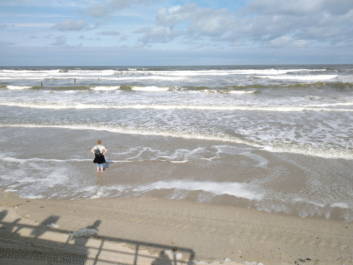 Sandstrand. Meer mit Gischt und mehreren Wellen-Bergen. Eine Person steht mit den Füßen am Rand vom Meer und blickt in die Ferne.