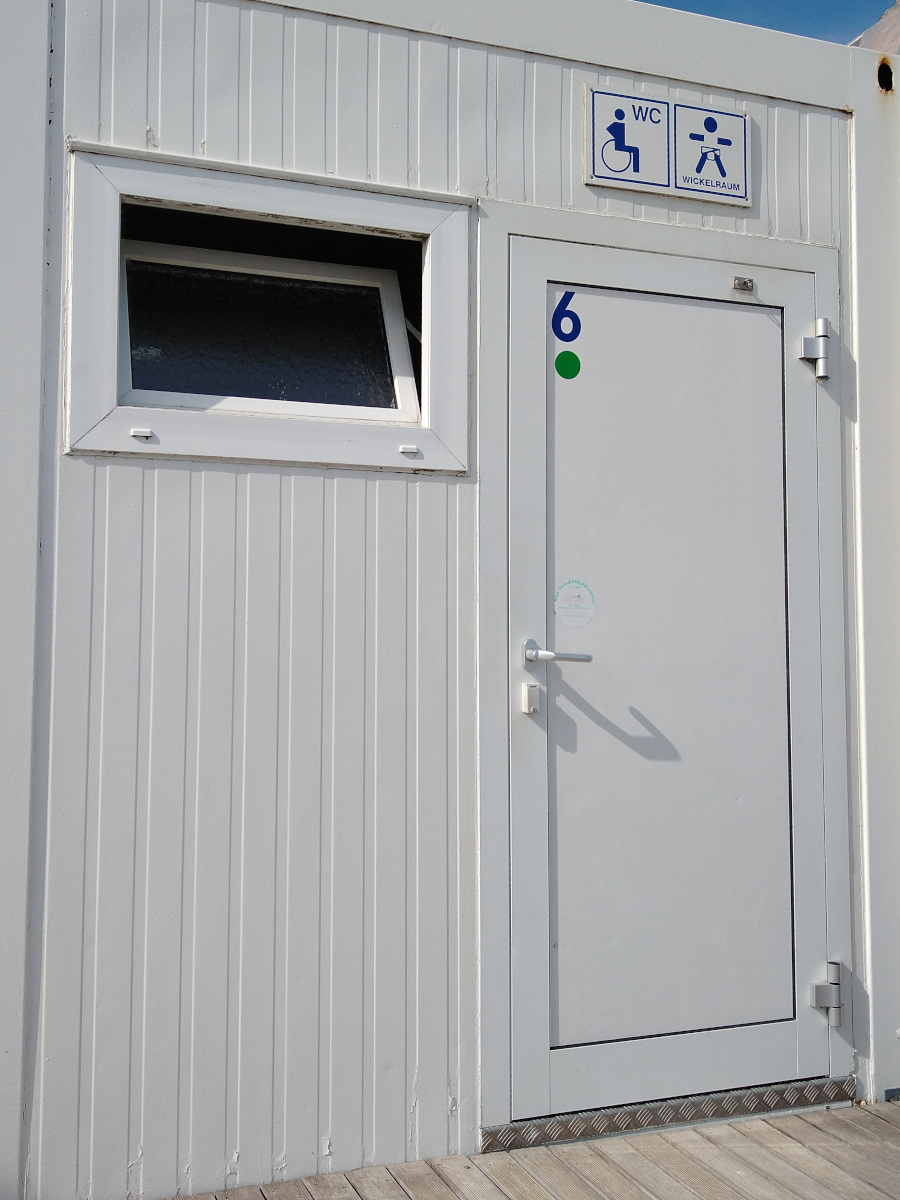 Weißer Container mit einer Tür. Über der Tür ein Schild mit Rollstuhlfahrer-Symbol und dem Text "WC". Und daneben ein Schild mit dem Text "Wickelraum".