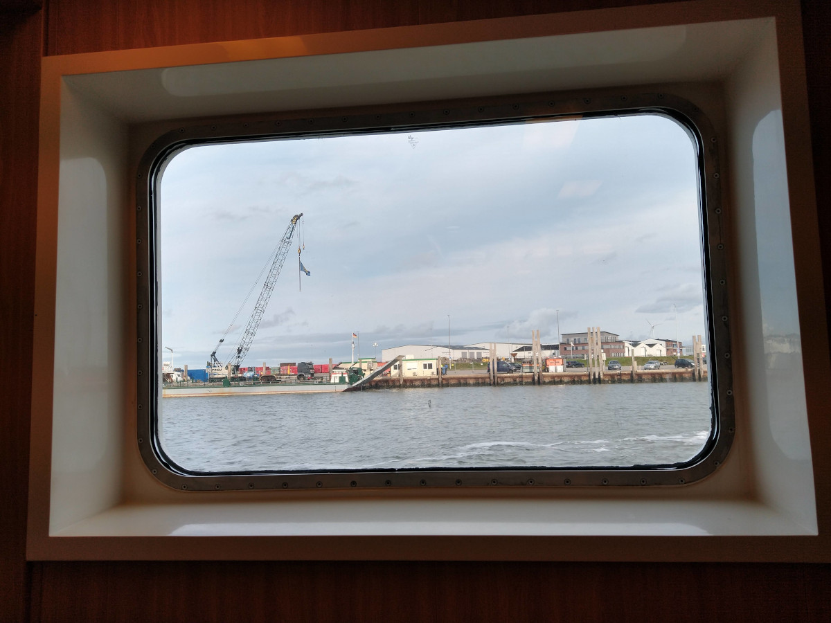 Blick aus dem Schiff auf eine Hafenanlage mit einem langen Kai und mehreren Industrie-Hallen.