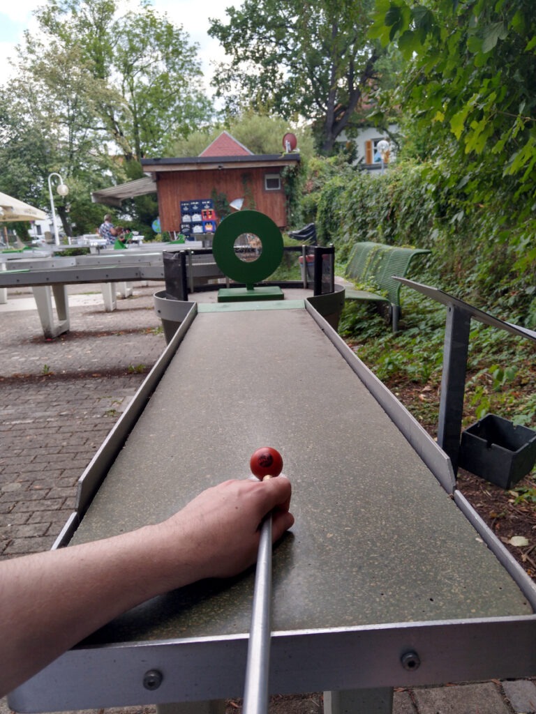 Blick vom Start der Pit-Pat-Bahn. Eine Kugel liegt auf dem Startpunkt, die linke Hand hält den Queue stoßbereit. Im Hintergrund befindet sich als Hindernis eine runde Scheibe mit einem Loch in der Mitte.