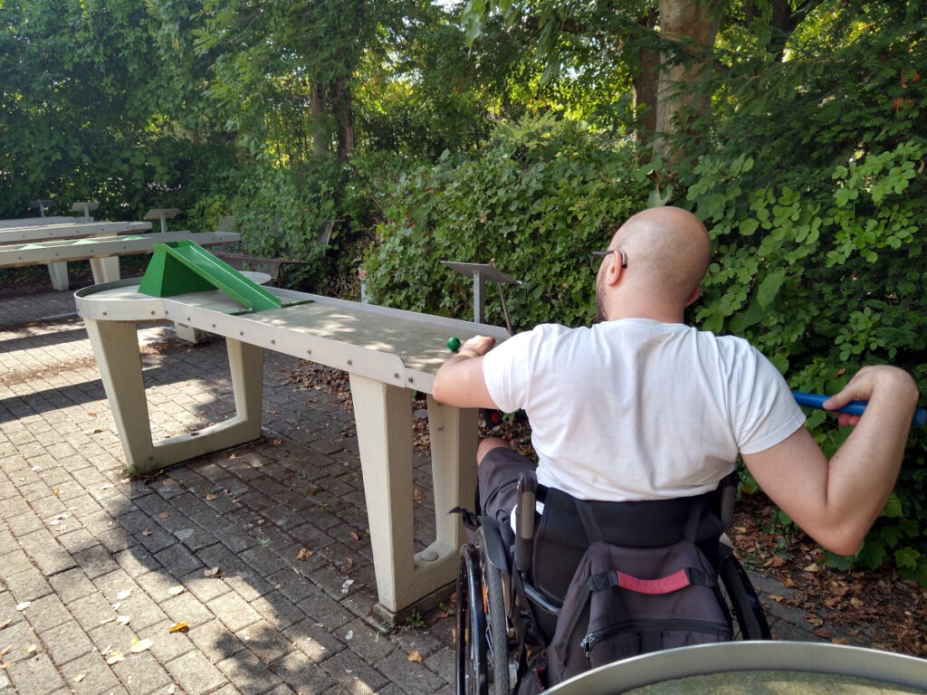 Rollstuhlfahrer an einer Pit-Pat-Bahn, der die Kugel auf dem Startpunkt mit dem Queue anvisiert. Auf der Bahn befindet sich als Hindernis eine mittige Rampe, die zum Loch führt.