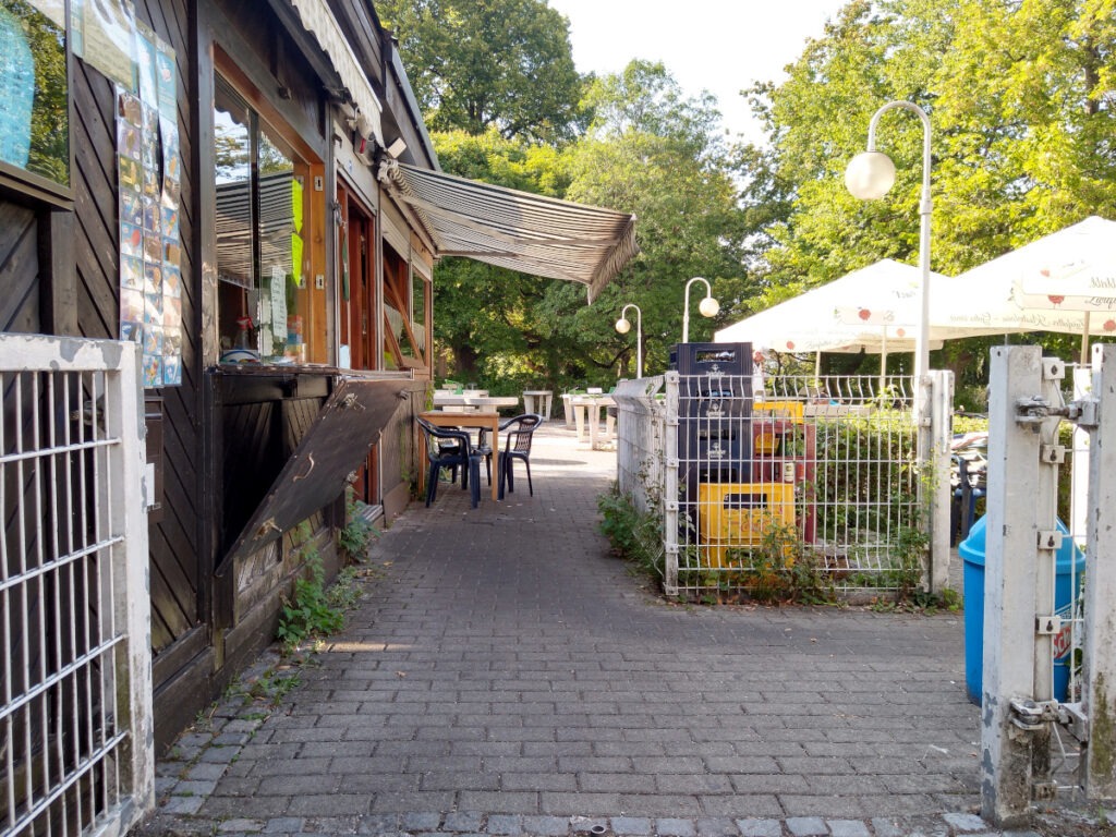 Links ein rustikales Imbissgebäude aus Holz. Geradeaus führt ein Durchgang zur Pit-Pat-Anlage, rechts geht es zum eingezäunten Sitzbereich des Imbisses.