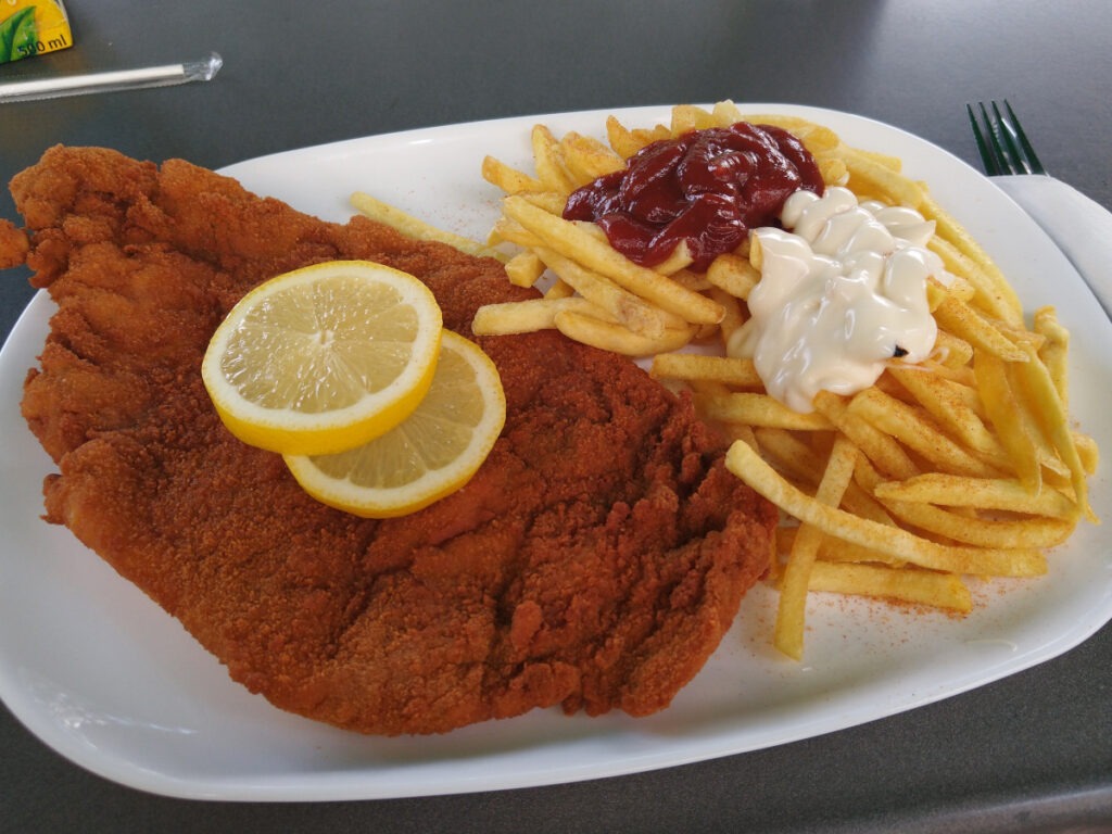 Teller mit großem Schnitzel mit goldbrauner Panade. Daneben Pommes frites mit Mayonnaise und Ketchup.