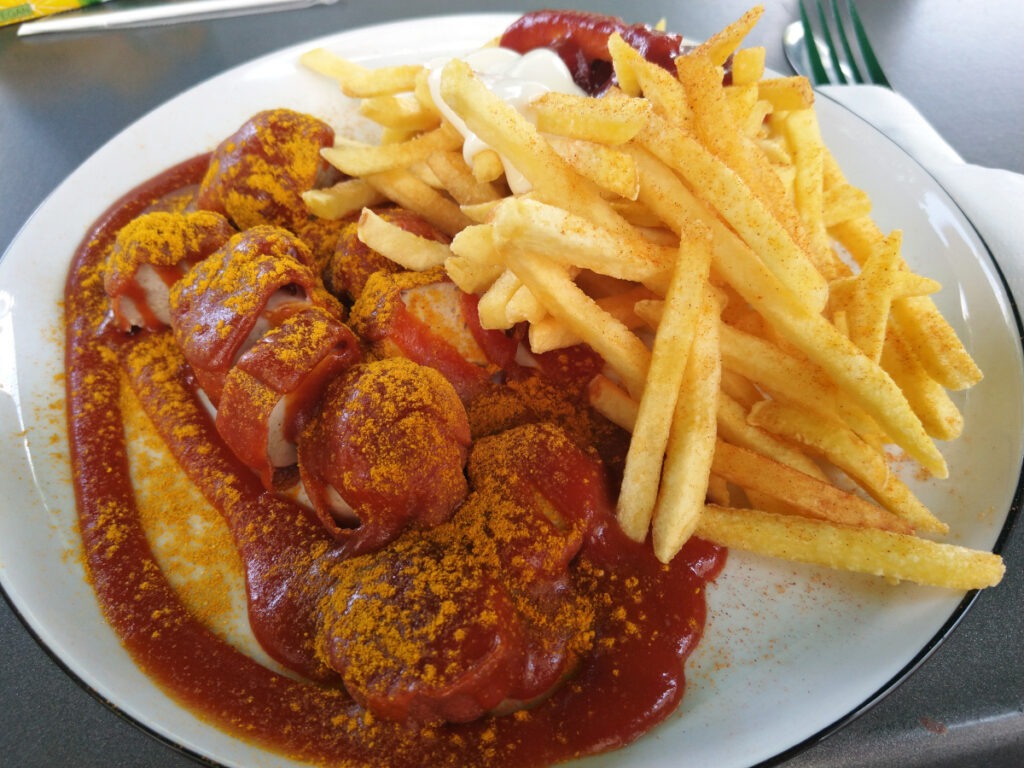 Teller mit links in Stücke geschnittener Currywurst mit reichlich Currysoße. Daneben eine Portion Pommes frites sowie separat Mayonnaise und Ketchup.