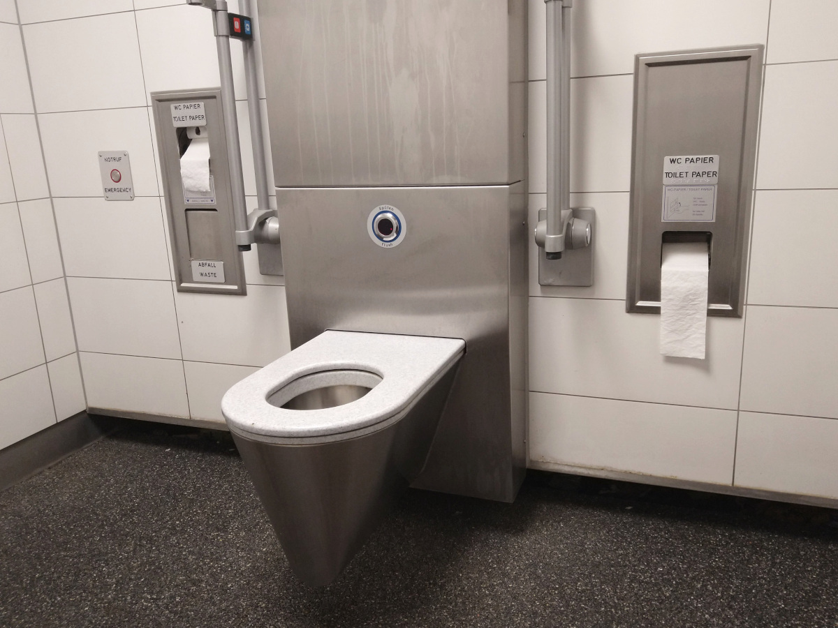 Behinderten-Toilette mit 2 Klapp-Griffen. Toilette aus Edelstahl. Boden aus Asphalt.