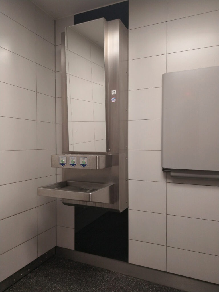 Metallwaschbecken mit schräg angebrachtem Spiegel. Über dem Waschbecken befinden sich Bedienknöpfe für Wasser, Seife und Föhn.