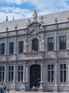 Zweistöckiges Gebäude mit hohen Fenstern und Säulen, über der Tür ein kleiner Balkon mit kunstvollen Statuen, darüber am Dachsimse weitere dekorative Figuren.