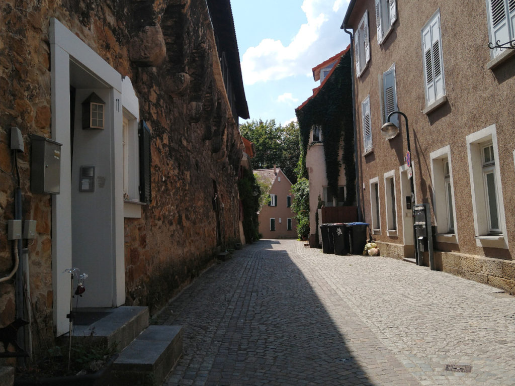 Weg mit unebenem Kopfstein-Pflaster. Links die alte Stadt-Mauer, in der sich immer wieder Eingangs-Türen zu Wohnungen befinden. Rechts moderne Häuser.