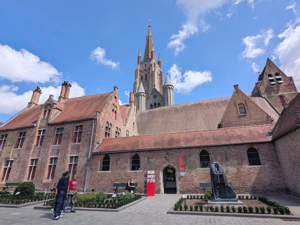Links zweistöckiges rotes Backsteingebäude, mittig überdachter gemauerter Gang, dahinter weitere rote Backsteingebäude, im Hintergrund mittig ein gemauerter Kirchturm.