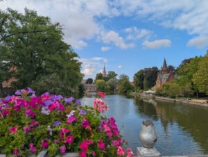 Rechteckiger See, umrahmt von Bäumen, rechts am Ufer ein kleines rotes Backsteinschlösschen, im Hintergrund eine Turmspitze, zentral unten im Bild ein Metallgeländer mit Blumenkasten und bunten Blumen.