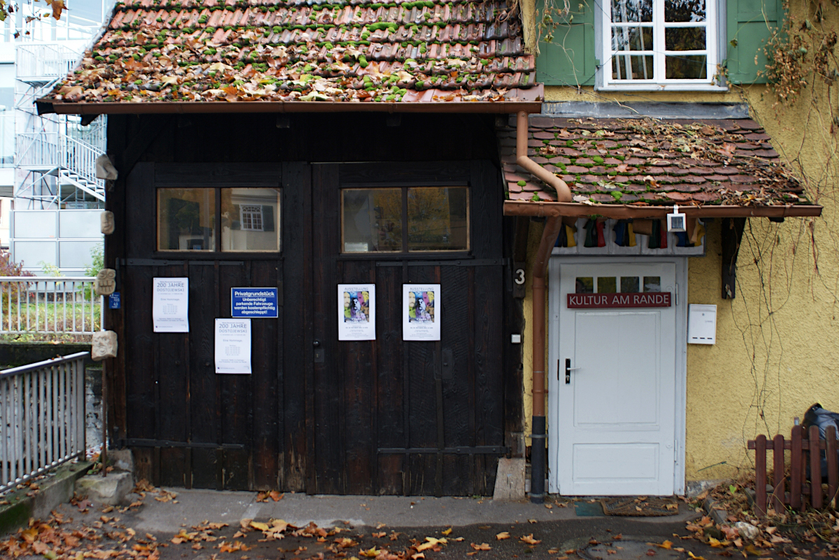 Außenansicht der "Alten Spinnerei". Links eine Garage mit 2-flügligem Holztor. Rechts kleines gelbes Häuschen.