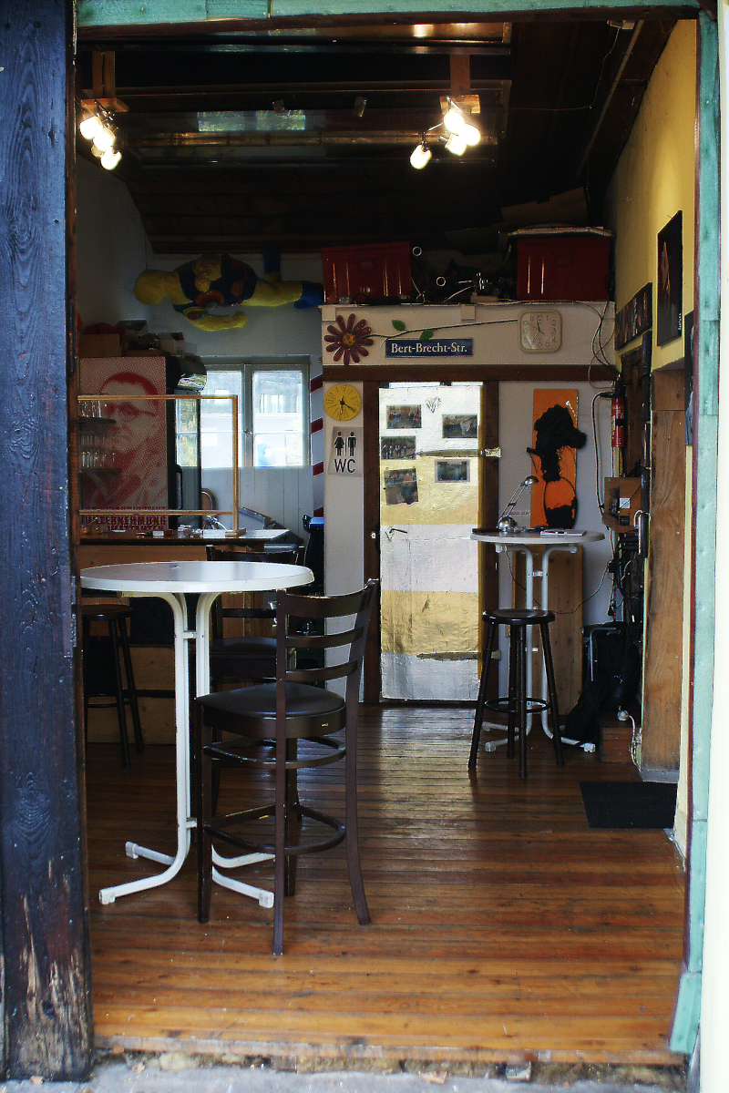 Blick in den Innenbereich mit dem Barbereich. Zwei Stehtische vorne und rechts. Links hinten eine Bar.