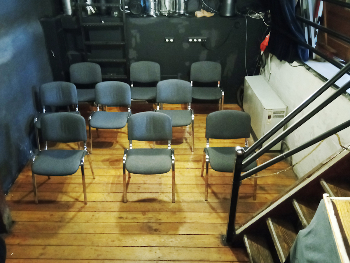 Blick von der Bühne in den Publikumsbereich. Im Publikumsbereich 3 Reihen mit je 3 Stühlen.