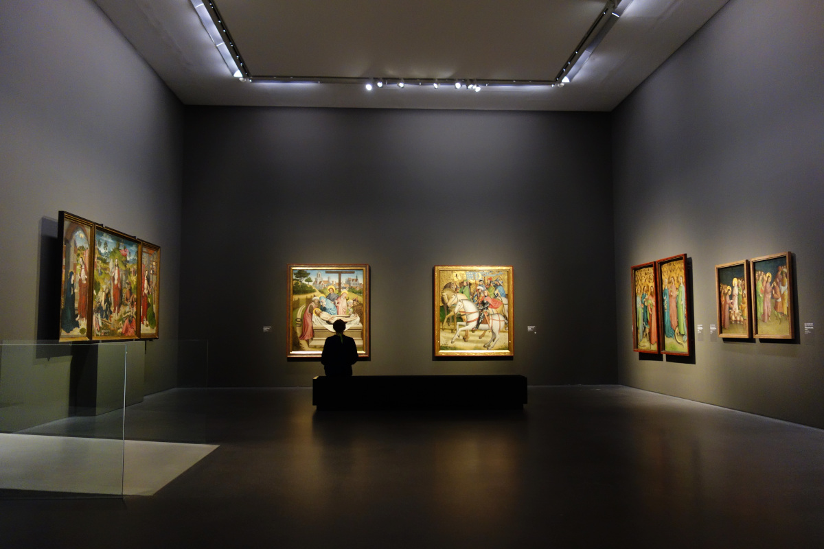 Saal mit mehreren Gemälden. Zentral sitzt eine Person auf einer Bank und betrachtet die Bilder