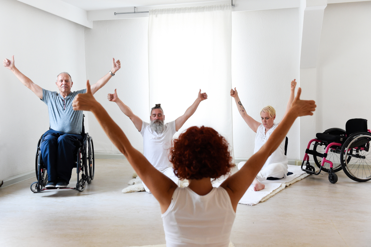 Yogagruppe mit einer Rollstuhlfahrer auf dem Boden und einem Rollstuhlfahrer im Rollstuhl