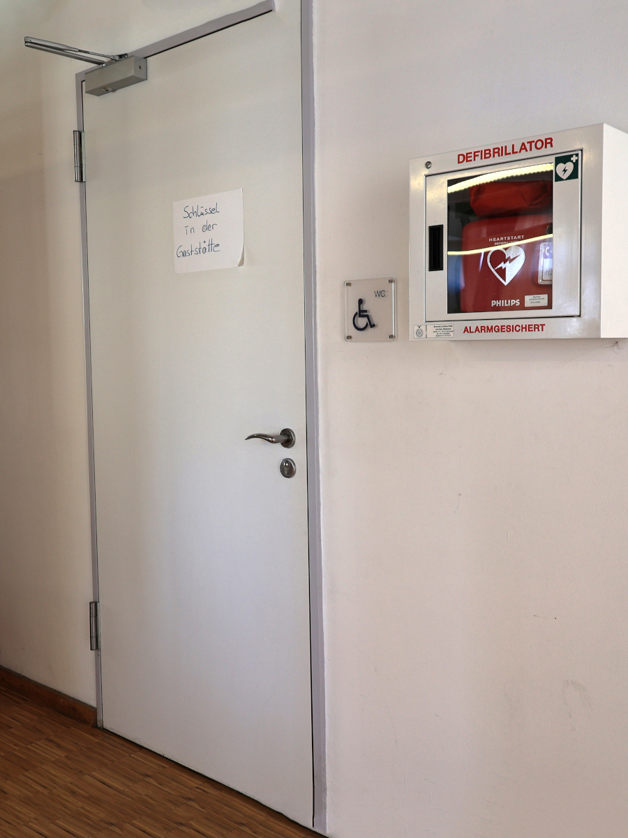 Tür von der Behinderten-Toilette. Auf einem Zettel an der Tür steht: "Schlüssel in der Gaststätte." Daneben an der Wand ein Defibrillator.